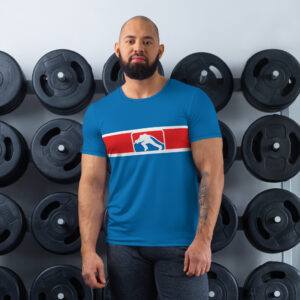 A man stands confidently in a gym setting, wearing a blue t-shirt with a red and white stripe across the chest, featuring the FCL (Firefighter Challenge League) logo in the center. The background includes weight plates stacked on racks, highlighting an active and athletic environment.