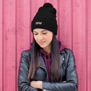 A woman with long dark hair is standing in front of a pink wooden wall, wearing a black pom-pom beanie with the UPW logo embroidered on the front. She is dressed in a leather jacket with a burgundy hoodie underneath.