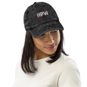 A woman wearing a black denim cap featuring the UPW logo embroidered in white and red on the front. The hat has a worn, stylish look, and the woman wears it with a relaxed expression.